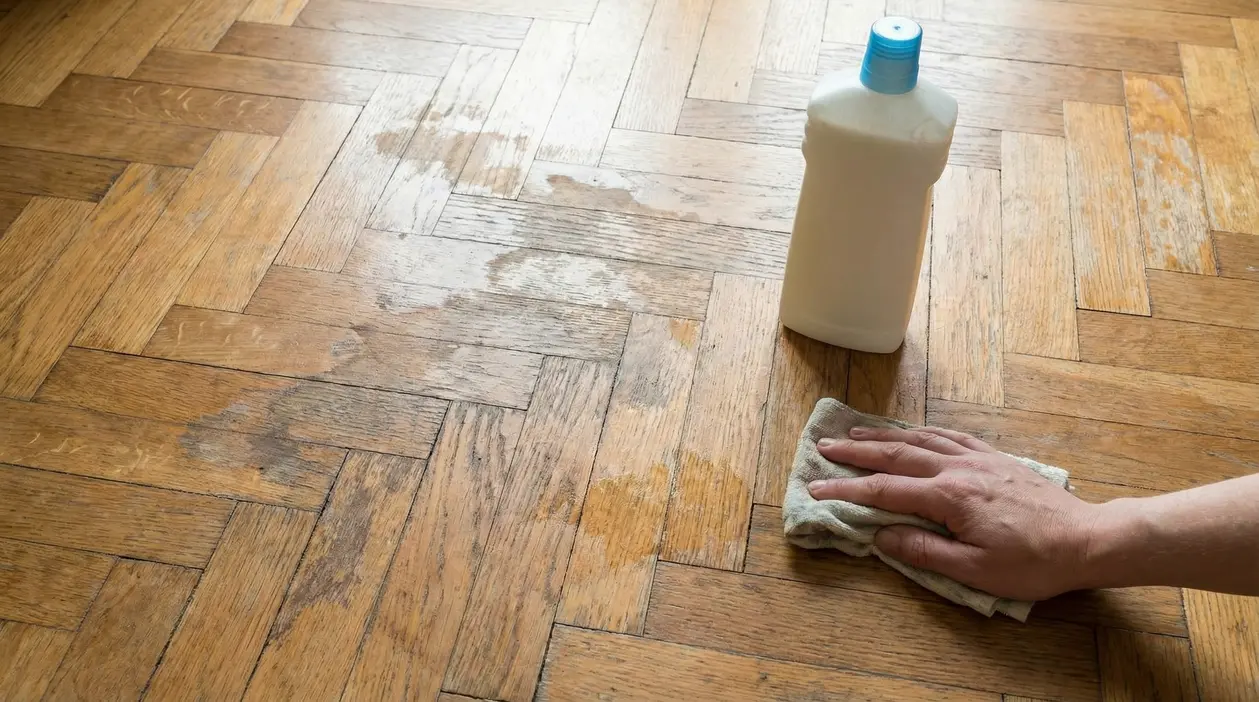 Mano che pulisce un parquet rovinato con un panno accanto a un flacone di detergente